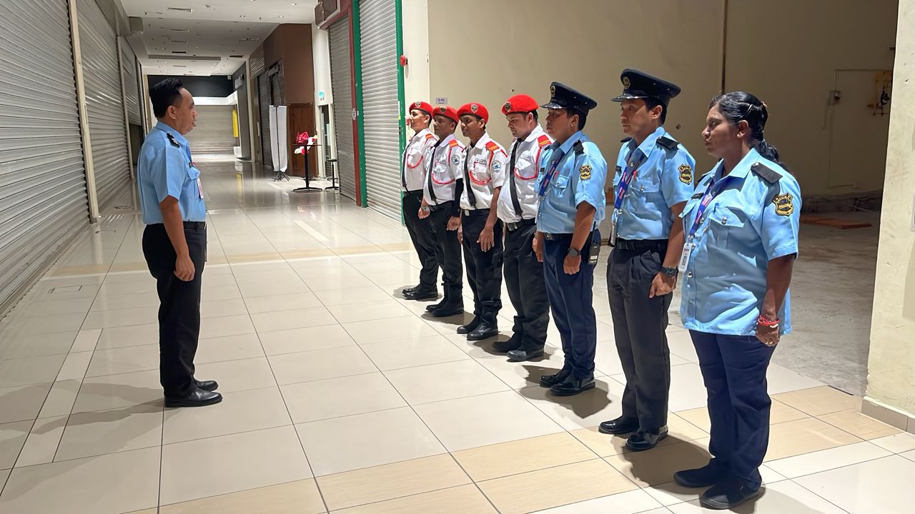 KSD Security guards in light-blue and white uniforms standing in formation for a supervisor inspection along a shopping mall corridor.