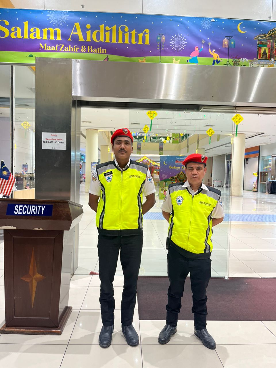 Two KSD Security guards in hi-vis vests and red berets standing guard at a mall entrance decorated with a Salam Aidilfitri banner.