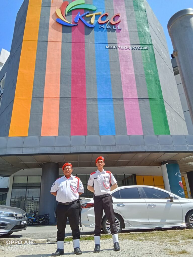 Two KSD Security guards in white shirts and red berets standing on duty outside the colourful KTCC Mall facade in Kuala Terengganu.
