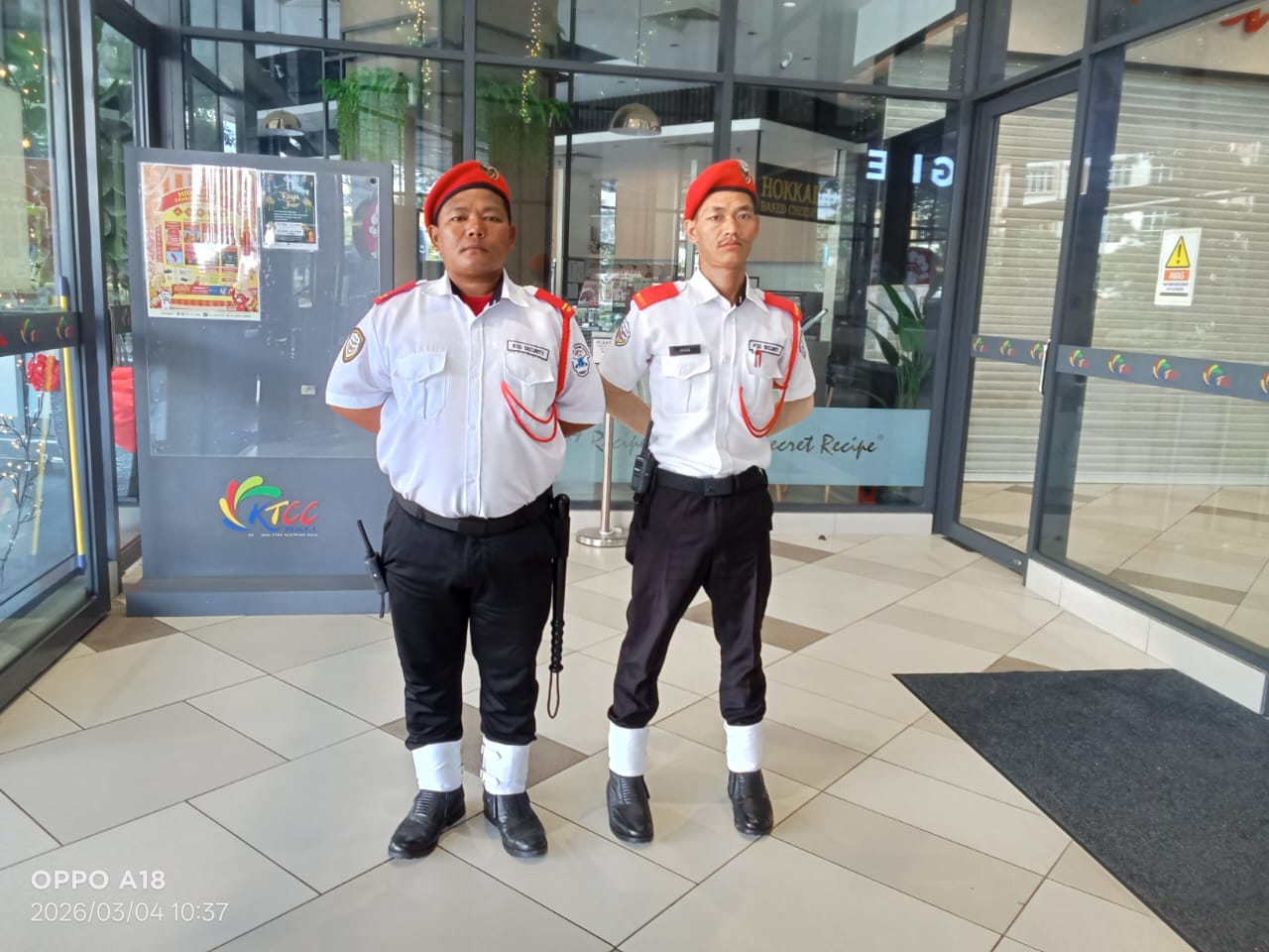 Two KSD Security guards in white shirts and red berets standing at attention inside the KTCC Mall lobby entrance.