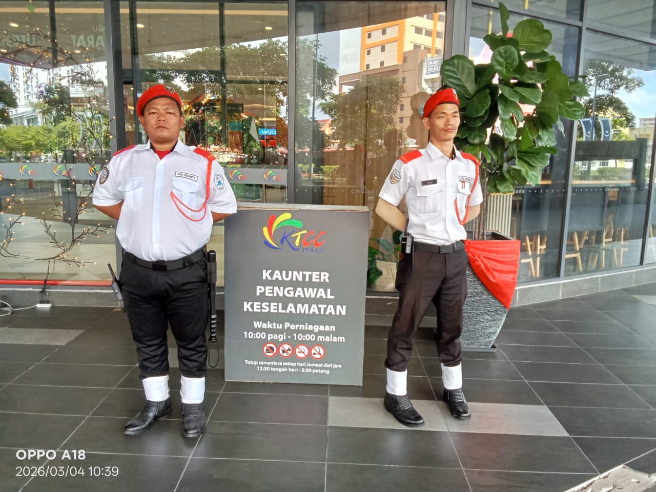 Two KSD Security guards on duty beside the KTCC Mall security counter signboard listing guard service hours.