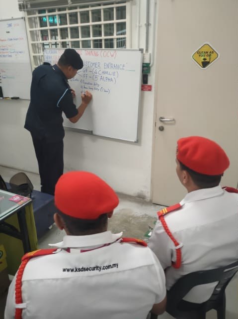 KSD Security trainer writing the Customer Entrance lesson on a whiteboard during an OCW class at AEON Falim while two uniformed KSD guards observe